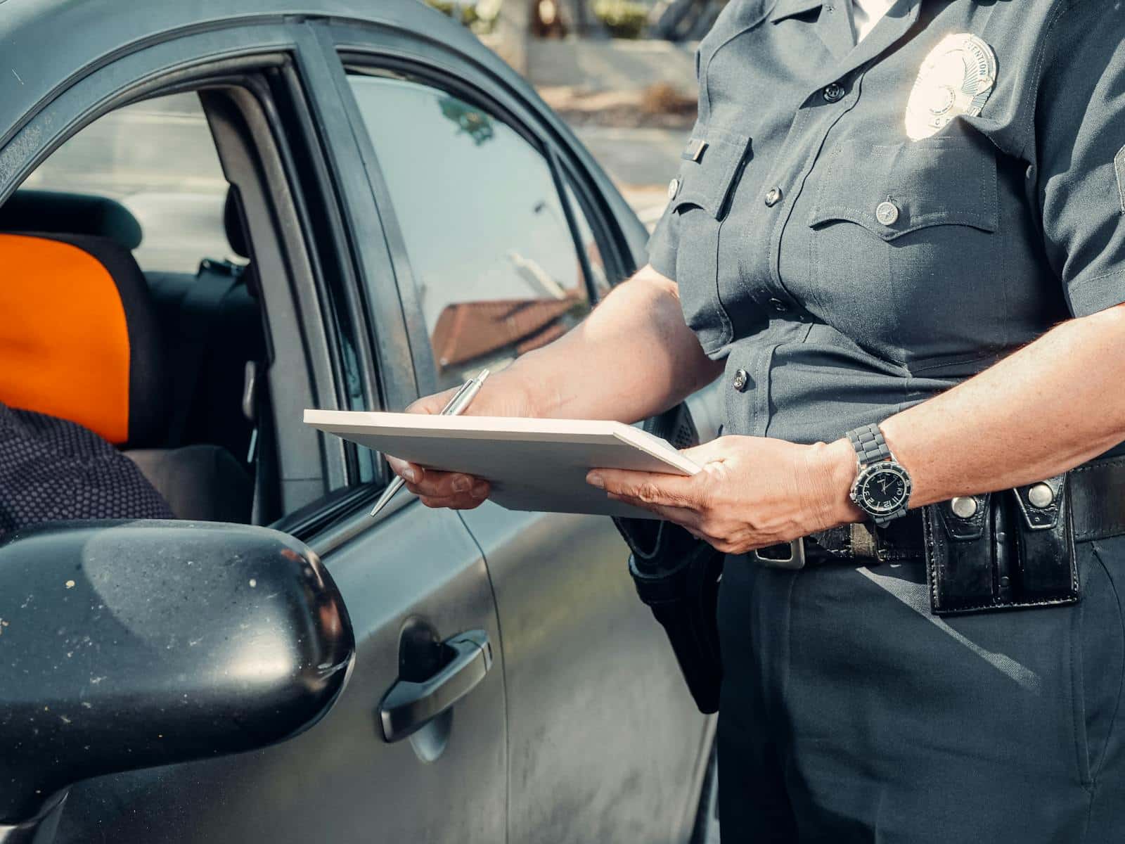 A police officer in uniform writes a ticket to a driver through the car window on a sunny day.